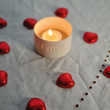 Load image into Gallery viewer, Looking down on a creamy white porcelain tealight holder with the word Love etched into it. A glowing candle and chocolate hearts.
