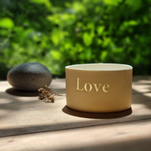 Load image into Gallery viewer, Creamy white porcelain tealight holder with the word Love etched into it.On a table bathed in natural light.