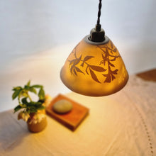 Load image into Gallery viewer, a warmly lit porcelain Lampshade etched with wild roses and a butterfly. A vase with flowers, a book and a pebble in the background.