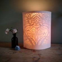 Load image into Gallery viewer, Porcelain lamp with symetrical entwined tree and birds in silhouette. Glowing warm orange.
