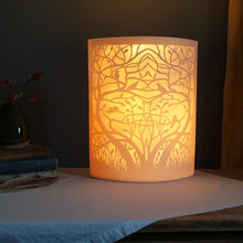 Load image into Gallery viewer, Porcelain lamp with symetrical entwined tree and birds in silhouette. Glowing warm orange.