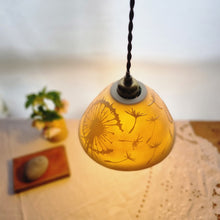 Load image into Gallery viewer, Lampshade glowing warmly with dandelion clocks and seeds etched onto it. Flowers, a book and a pebble on a table in the background.
