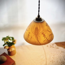 Load image into Gallery viewer, Lampshade glowing warmly, made of porcelain with thistles and teasels etched onto it. Flowers, a book and a pebble on a table in the background.
