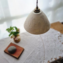 Load image into Gallery viewer, Lampshade warm cream unlit porcelain, with thistles and teasels  etched onto it. Flowers, a book and a pebble on a table in the background.
