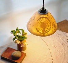 Load image into Gallery viewer, Lampshade glowing warmly with dandelion clocks and seeds etched onto it. Flowers, a book and a pebble on a table in the background.
