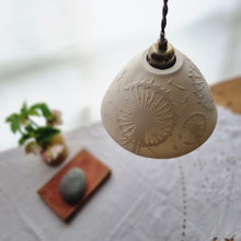 Load image into Gallery viewer, Lampshade warm cream unlit porcelain, with dandelion clocks and seeds etched onto it. Flowers, a book and a pebble on a table in the background.
