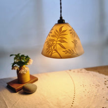 Load image into Gallery viewer, Porcelain Lampshade glowing gently, etched with swirling Ash leaf design. Yellow vase with flowers in the background.
