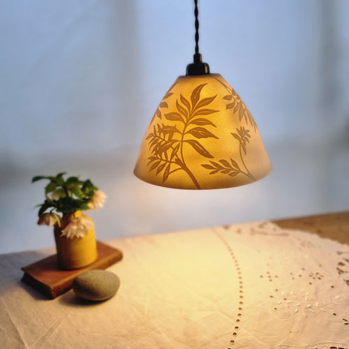 Porcelain Lampshade glowing gently, etched with swirling Ash leaf design. Yellow vase with flowers in the background.