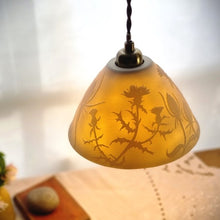 Load image into Gallery viewer, Lampshade glowing warmly with thistles and Teasels etched onto it. Flowers, a book and a pebble on a table in the background.
