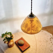Load image into Gallery viewer, Lampshade glowing warmly,  made of porcelain with thistles and teasels etched onto it. Flowers, a book and a pebble on a table in the background.

