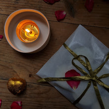 Load image into Gallery viewer, a lit tealight giving off a soft glow next to a wrapped present. 
