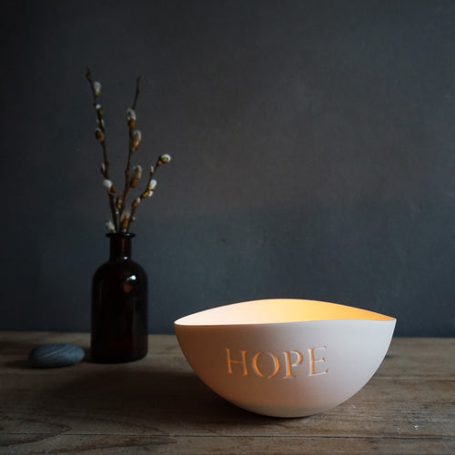 Porcelain bowl lit with a candle with HOPE etched into it, pebble and bottle with pussy willow on the left.