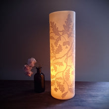 Load image into Gallery viewer, Porcelain cylinder Lamp etched with an Oak tree design in silhouette, glowing warmly.
